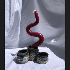 Puede incluir: Una vela roja ondulada se encuentra en un portavelas de cerámica con tres pequeñas tazas redondas de cerámica esmaltada en marrón, verde y blanco.