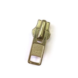 May include: A close-up of a gold-colored zipper pull on a white background. The zipper pull has a rectangular base with the letters "YKK" stamped on it. The pull is made of metal and has a shiny finish.