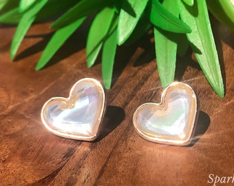 Könnte beinhalten: Ein Paar herzförmige Ohrringe mit perlmuttartigem Finish und goldenen Akzenten. Die Ohrringe liegen auf einer Holzoberfläche mit grünem Laub im Hintergrund. Sparkly Chic Jewels steht unter den Ohrringen.