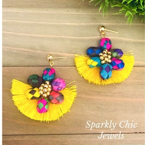 May include: A pair of yellow tassel earrings with a colorful woven flower design and gold accents. The earrings are on a wooden surface with green foliage in the background. "Sparkly Chic Jewels" is written in white text on the wooden surface.