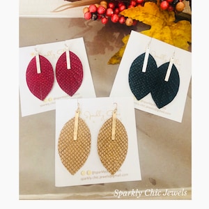 May include: Three pairs of leaf-shaped earrings in burgundy, navy, and tan, each with a textured finish and a vertical gold bar accent. The earrings are displayed on white cards with the text "Sparkly Chic Jewels".
