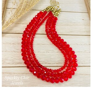 May include: A multi-strand necklace made of red crystal beads. The necklace has a gold clasp and is layered for a dramatic look.