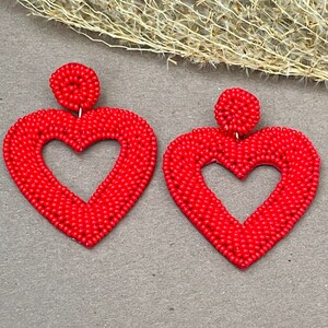 Pu&ograve; includere: Un paio di orecchini a forma di cuore rosso fatti di piccole perline. Gli orecchini sono grandi e pendenti.