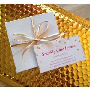 Pu&ograve; includere: Una scatola bianca con un nastro dorato legato intorno. La scatola &egrave; appoggiata su una busta di bolle dorata. Un biglietto da visita con il testo "Sparkly Chic Jewels" e il nome "Marytha Merrill" &egrave; visibile nell'immagine. Il biglietto da visita include anche l'indirizzo del sito web "sparklychicjewels@gmail.com", la pagina Facebook "www.facebook.com/sparklychicjewels" e il negozio Etsy "www.etsy.com/shop/sparklychicjewels".