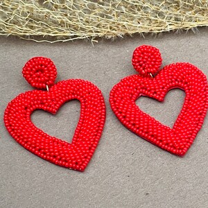 Pu&ograve; includere: Un paio di orecchini rossi a forma di cuore fatti di piccole perline. Gli orecchini sono pendenti e hanno un'apertura a forma di cuore al centro.