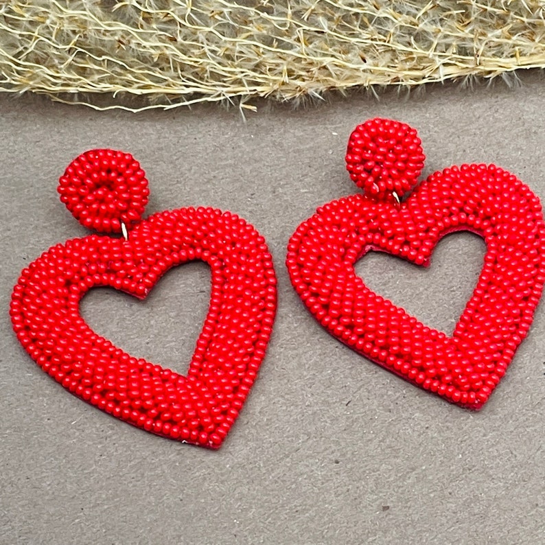 Pu&ograve; includere: Un paio di orecchini rossi a forma di cuore fatti di piccole perline. Gli orecchini sono pendenti e hanno un'apertura a forma di cuore al centro.