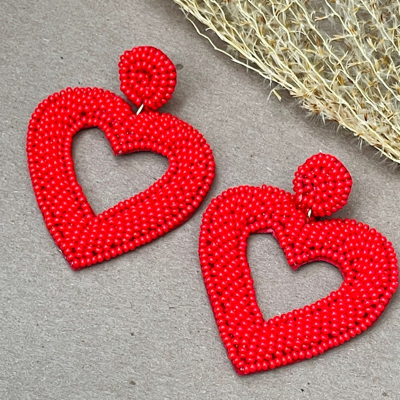 Pu&ograve; includere: Un paio di orecchini a forma di cuore rosso fatti di piccole perline. Gli orecchini sono grandi e pendono dall'orecchio.