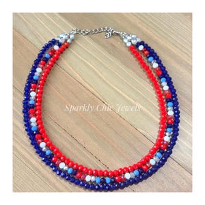 May include: A multi-strand beaded necklace featuring faceted beads in shades of red, dark blue, light blue, and white. The necklace includes a silver-toned metal chain extender and clasp. The text Sparkly Chic Jewels is visible in the center.