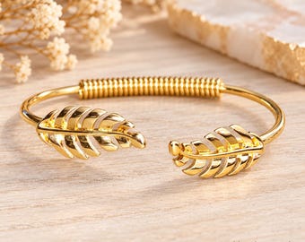 May include: A gold-toned open cuff bracelet featuring two symmetrical leaf-shaped ends and a central coiled wire section. The polished metal surface reflects light, creating a bright appearance. The design is circular with a gap between the leaves.
