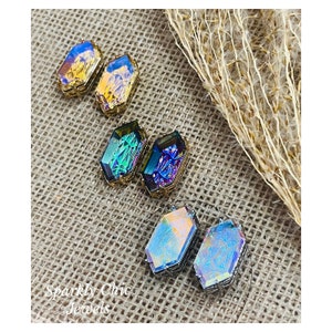 Può includere: Un set di sei orecchini in vetro iridescente in tre colori diversi: oro, blu e viola. Gli orecchini hanno la forma di ottagoni e una finitura metallica. Gli orecchini sono esposti su uno sfondo di juta.