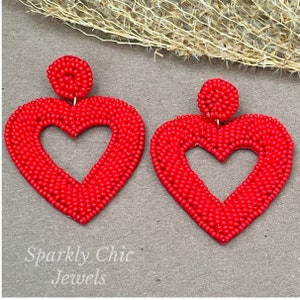 Pu&ograve; includere: Un paio di orecchini a forma di cuore rosso fatti di piccole perline. Gli orecchini hanno una forma di cuore cava al centro. Gli orecchini sono pendenti e hanno una piccola perla rotonda in cima.