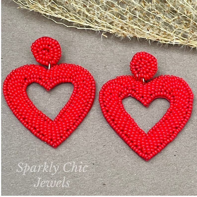 Pu&ograve; includere: Un paio di orecchini a forma di cuore rosso fatti di piccole perline. Gli orecchini hanno una forma di cuore cava al centro. Gli orecchini sono pendenti e hanno una piccola perla rotonda in cima.