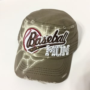 May include: A khaki green baseball cap with a distressed look. The cap has a white and brown baseball graphic with the word "Baseball" in brown script and the word "Mom" in white with rhinestones.