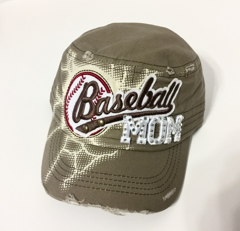 May include: A khaki green baseball cap with a distressed look. The cap has a white and brown baseball graphic with the word "Baseball" in brown script and the word "Mom" in white with rhinestones.