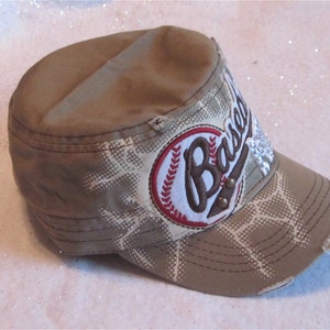 May include: A tan and white baseball cap with a distressed look. The cap has a red and white baseball patch with the word "Baseball" embroidered on it. The patch has rhinestones on it.