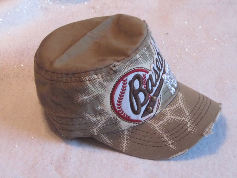 May include: A tan and white baseball cap with a distressed look. The cap has a red and white baseball patch with the word "Baseball" embroidered on it. The patch has rhinestones on it.
