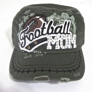 May include: A green camouflage baseball cap with a distressed look. The cap has a white patch with the words "Football Mom" in black lettering and rhinestones. A brown football graphic is on the patch.
