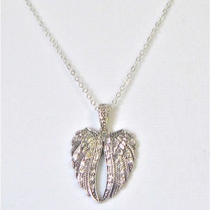 Puede incluir: Un collar de plata con un colgante en forma de coraz&oacute;n que presenta dos alas de &aacute;ngel adornadas con brillantes diamantes de imitaci&oacute;n.