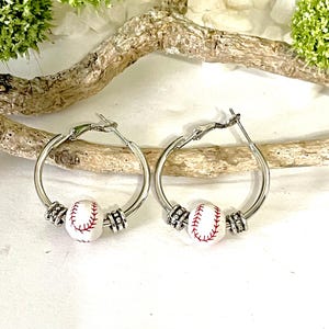 May include: Silver hoop earrings with a white baseball charm. The baseball charm has red stitching.
