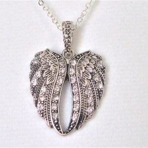 Puede incluir: Collar plateado con un colgante en forma de coraz&oacute;n con alas de &aacute;ngel adornadas con brillantes transparentes.