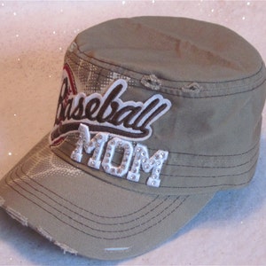 May include: A khaki green baseball cap with a distressed look. The cap has a white patch with the words "Baseball Mom" in brown and white lettering. The word "Mom" is embellished with rhinestones.