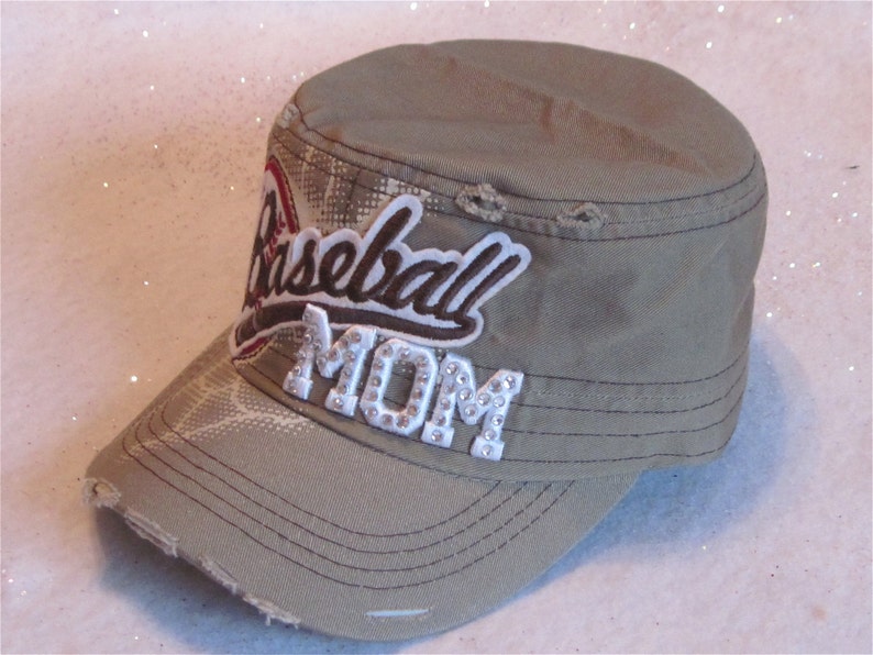 May include: A khaki green baseball cap with a distressed look. The cap has a white patch with the words "Baseball Mom" in brown and white lettering. The word "Mom" is embellished with rhinestones.