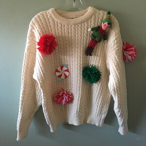 May include: Cream-colored cable knit sweater decorated with red, white, and green pom-poms, a candy-shaped applique, and a small elf doll. The sweater is hanging on a clear hanger against a light green wall.