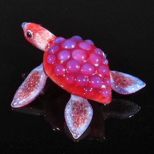 May include: A small glass turtle sculpture with a red and purple shell. The shell is covered in small, round, purple bubbles.