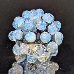 May include: A cluster of light blue, spherical glass beads with a speckled, opalescent appearance. The beads are arranged in a rounded shape, reflecting light and creating a shimmering effect. The background is black.