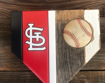 Puede incluir: Un home plate de béisbol de madera con una sección pintada de rojo, blanco y negro que presenta el logotipo de los St. Louis Cardinals. Una pelota de béisbol blanca está ubicada en la madera.
