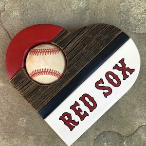 May include: A wooden heart-shaped sign with a baseball in the center. The sign is painted in red, white, and blue with the words "RED SOX" in red letters.