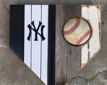 May include: A wooden baseball home plate with a white and black striped background and the letters "NY" in black. A baseball is set into the home plate.