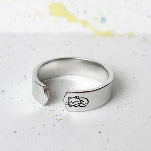 May include: A silver ring with a cat design engraved on the band. The ring is open on one side, making it adjustable.