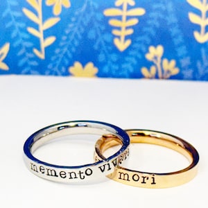 Op de afbeelding: Twee ringen, één zilver en één goud, in elkaar verweven. De zilveren ring heeft de woorden "memento vivere" en de gouden ring "mori" gegraveerd. De achtergrond is blauw met gouden bloemmotieven.