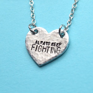 Może przedstawiać: Srebrny naszyjnik z sercowatym wisiorkiem z napisem "Always Keep Fighting".