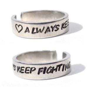 May include: Two silver rings with engraved messages. The top ring says "♡ ALWAYS KEEP" and the bottom ring says "S KEEP FIGHTING".
