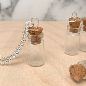 May include: A silver chain necklace with a small glass bottle pendant and a cork stopper. The bottle is clear and empty. There are several other similar bottles in the background.