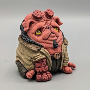 Puede incluir: Figura de pug rojo con ojos amarillos, cuernos y perilla negra. El pug lleva una chaqueta color canela y pantalones oscuros. La figura tiene un diseño único y estilizado.
