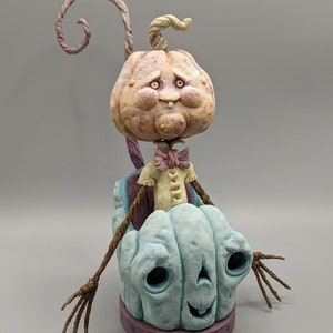 May include: A whimsical pumpkin figurine with a blue pumpkin body and a pale pink pumpkin head. The figurine has a long, twisted stem and two twig arms. The pumpkin head has a wide smile and large eyes.