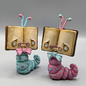 May include: Two bookworm figurines with open books for heads, wearing glasses and bow ties. One is blue with pink polka dots and the other is pink with blue polka dots.