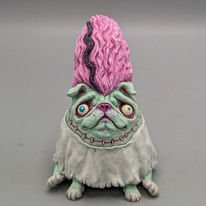 May include: A unique, handcrafted figurine of a pug dog with a Frankenstein's monster theme. The pug has a green face, stitched neck, and a pink, textured hairstyle with a black stripe. It wears a white shroud.