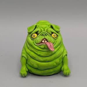 Könnte beinhalten: Eine grüne, mopsartige Skulptur mit groteskem Aussehen. Die Skulptur hat gelbe Augen, eine rosa Zunge und eine faltige Textur. Der Mops sitzt, wobei seine Vorderpfoten sichtbar sind. Das Gesamtdesign ist cartoonhaft und leicht beunruhigend.