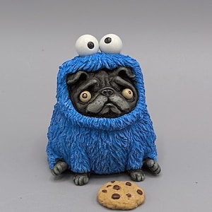 Könnte beinhalten: Figur einer schwarzen Mops-Figur in einem blauen, pelzigen Kostüm mit großen weißen Augen, das einem bekannten Charakter ähnelt. Ein kleiner, runder Keks mit Schokoladenstückchen liegt vor dem Mops. Die Figur steht auf einer grauen Oberfläche.