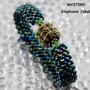 Op de afbeelding: Een kralenarmband met een groene, blauwe en gouden kleurstelling. De armband heeft een centrale kraal die groter is dan de rest van de kralen. De armband heet "MYSTERY" van Stephanie Jakob.