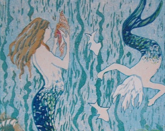 May include: A print titled "Mermaids" by Ruke Heier, featuring two mermaids in a turquoise ocean scene. One mermaid holds a seashell, while the other swims nearby. The artwork includes fish and aquatic plants.