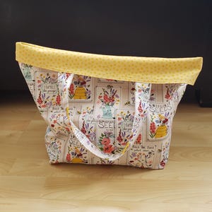 May include: A white tote bag with a yellow polka dot top. The bag has a floral print with the words "Humble Bee Garage", "Wildflower Seeds", and "Feed The Bees" on it.