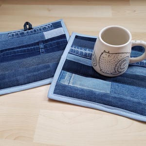 May include: Two denim pot holders with light blue trim and a white mug with a cat illustration. The pot holders are made from repurposed denim patches. The mug is sitting on one of the pot holders.