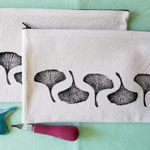 May include: Two white canvas zippered pouches with black ginkgo leaf prints. The pouch on the bottom has five leaves, while the pouch on top has one leaf. A teal blue ginkgo leaf carving tool is next to the pouches.