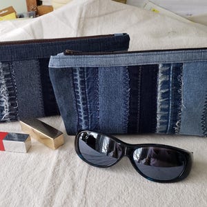 Peut inclure: Deux pochettes zippées en jean, l'une avec un motif patchwork et l'autre avec un motif en jean uni. Une paire de lunettes de soleil noires et deux tubes de rouge à lèvres sont sur une surface blanche.