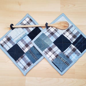 May include: Two square pot holders with a patchwork design of denim and plaid fabrics in shades of blue, white, and brown. A wooden spoon rests across the top of the pot holders. The pot holders have a light blue border.